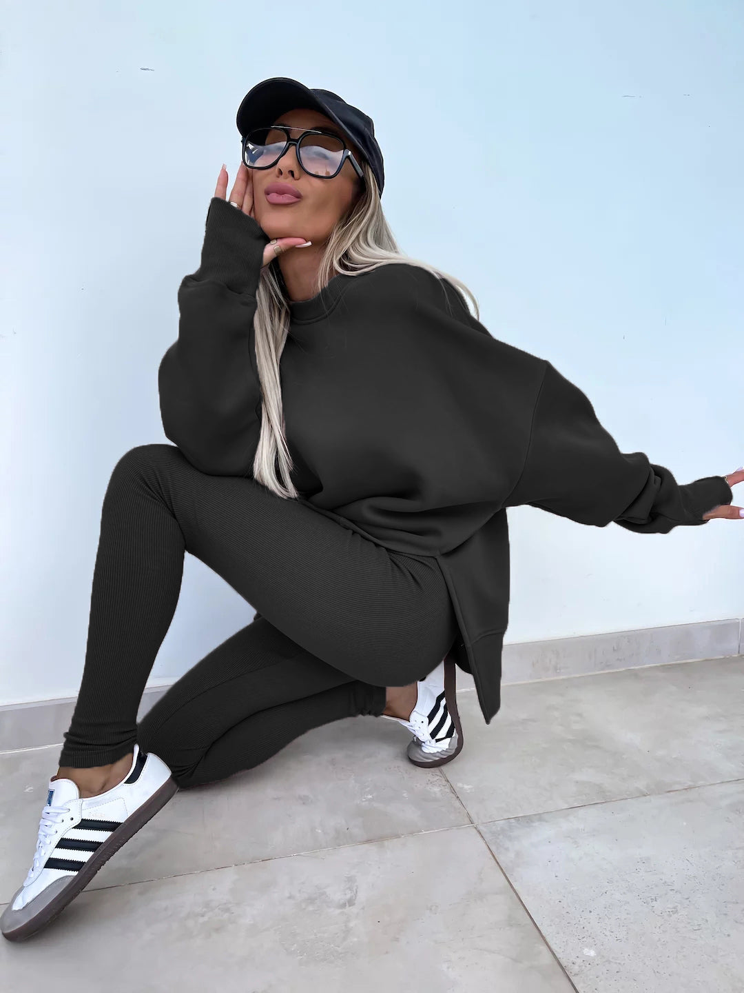 Black round-neck loose sweatshirt with long hip-covering length and side slits at the hem, paired with high-elasticity slim-fit trousers. Two-piece set. Model shows in a side-leaning and semi-squatting pose.