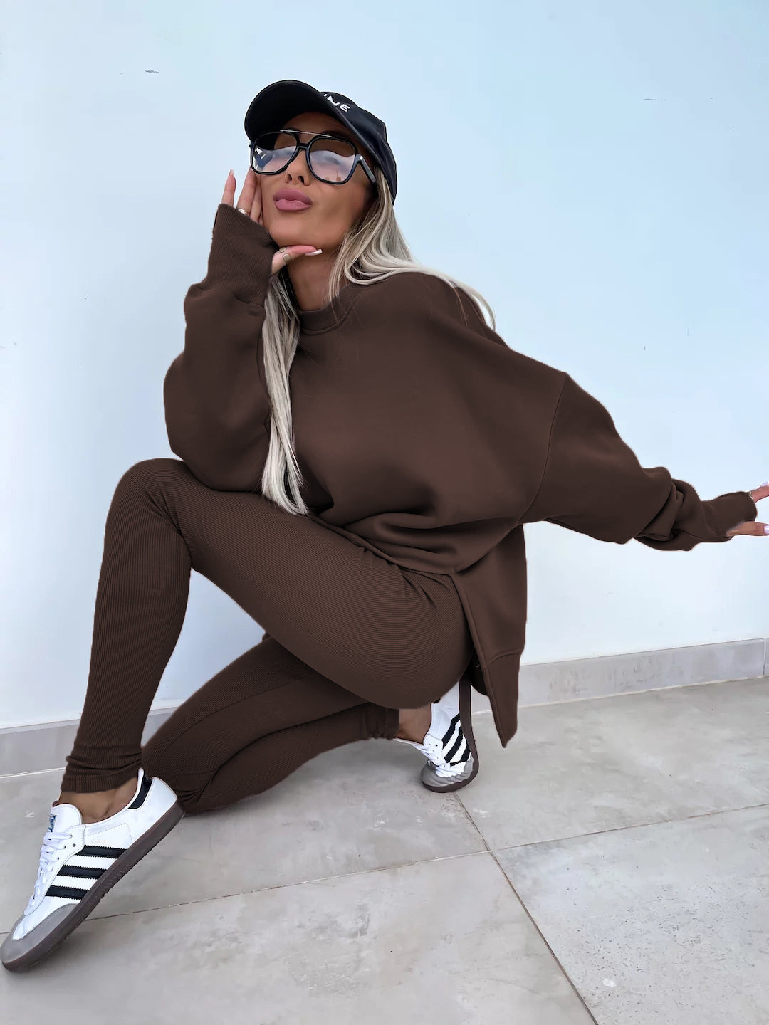 Coffee round-neck loose sweatshirt with long hip-covering length and side slits at the hem, paired with high-elasticity slim-fit trousers. Two-piece set. Model shows in a side-leaning and semi-squatting pose.
