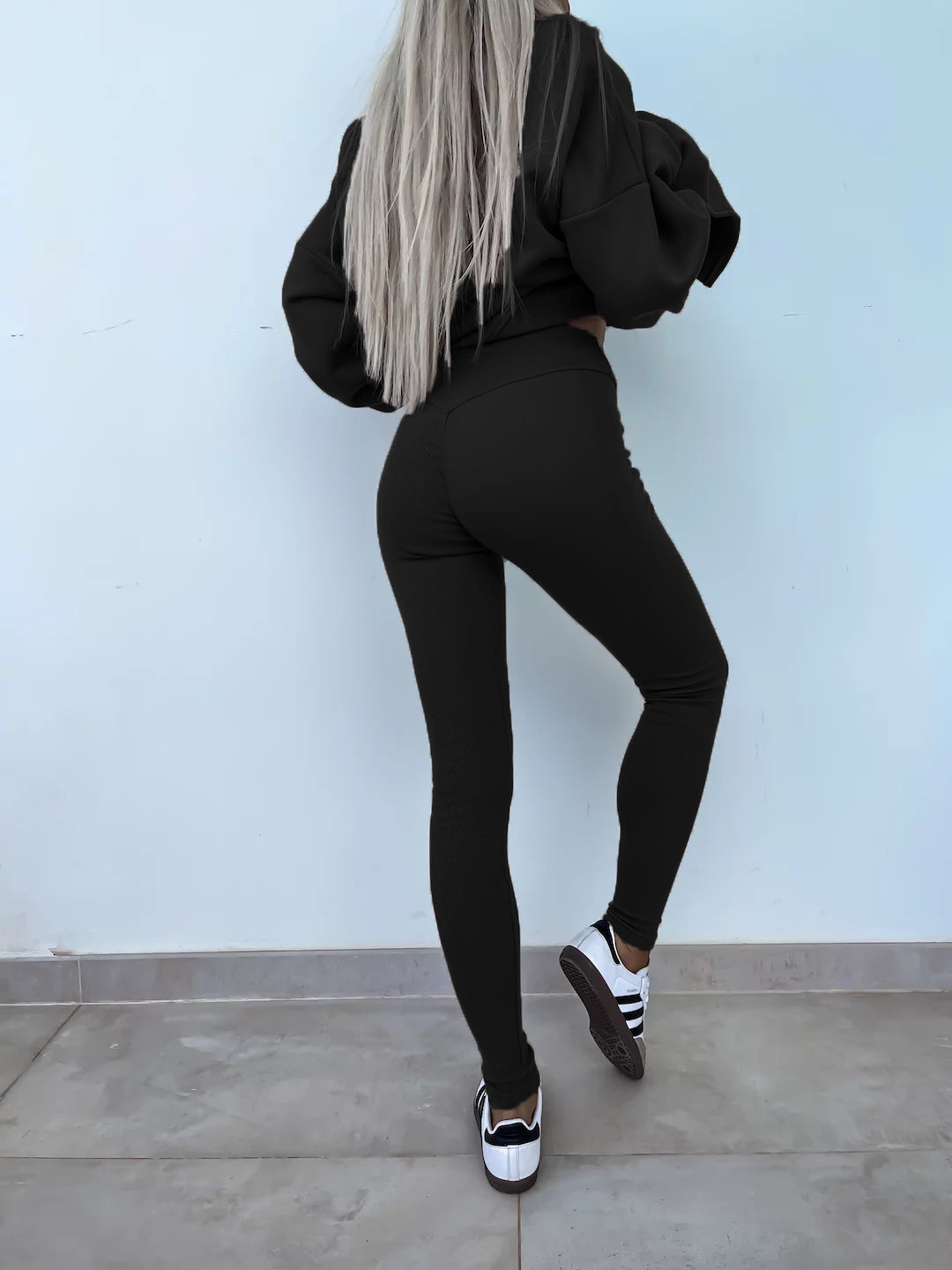 Black round-neck loose sweatshirt with long hip-covering length and side slits at the hem, paired with high-elasticity slim-fit trousers. Leisure suit. Model is wearing and showing the back.